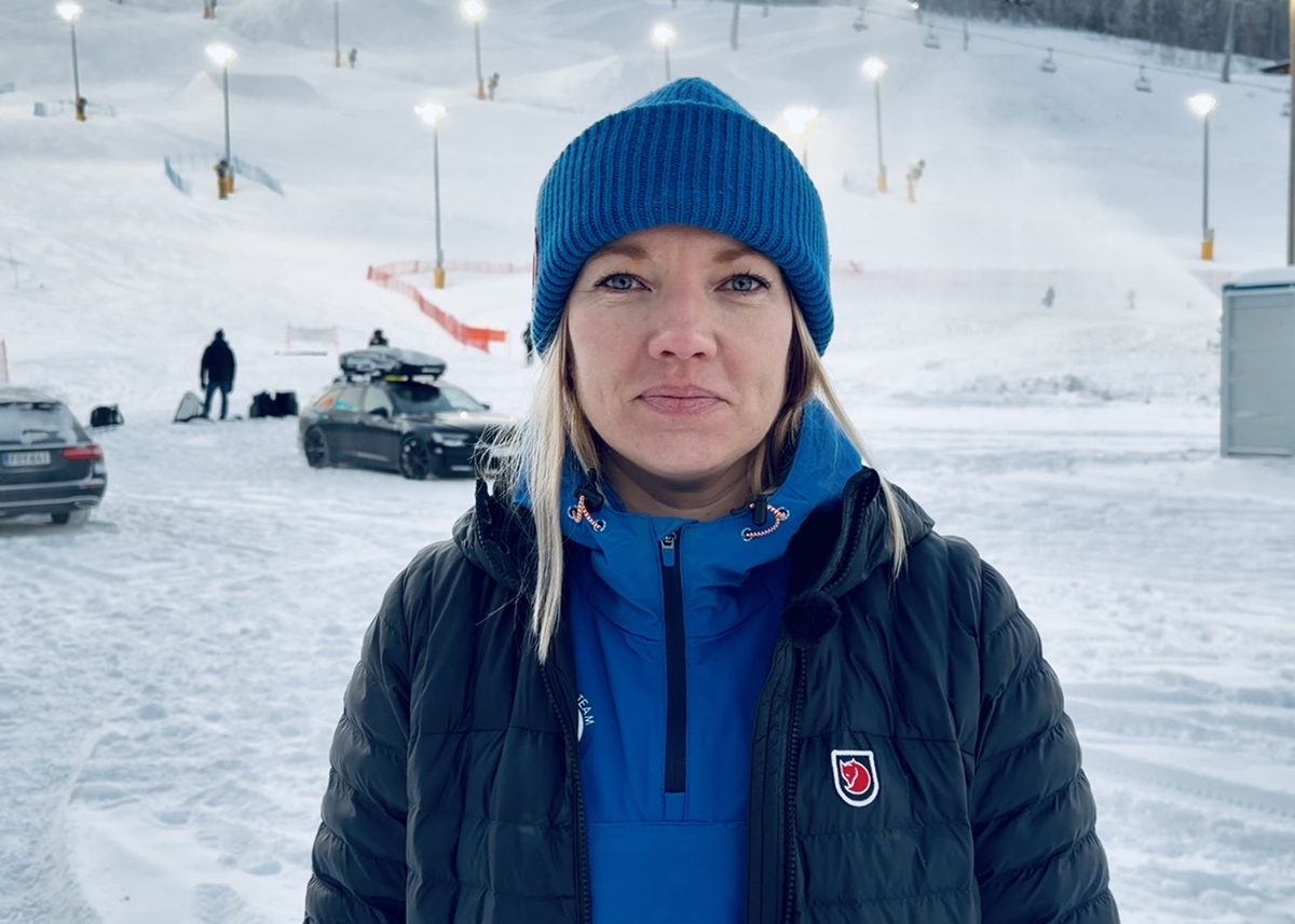 Vaaleahiuksinen Eveliina hymyilee katsoen suoraan kameraan sinisessä pipossa,  laskettelurinne taustallaan. 